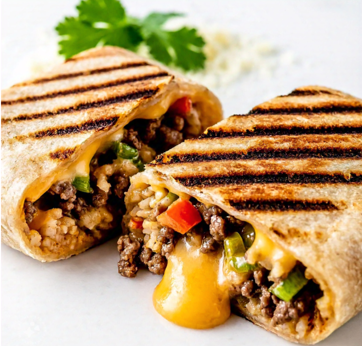 A close-up shot of a grilled cheese burrito cut in half to reveal its filling, including gooey melted cheese, seasoned beef, fluffy rice, and colorful veggies. The backdrop includes a sprinkle of grated cheese and fresh cilantro leaves.