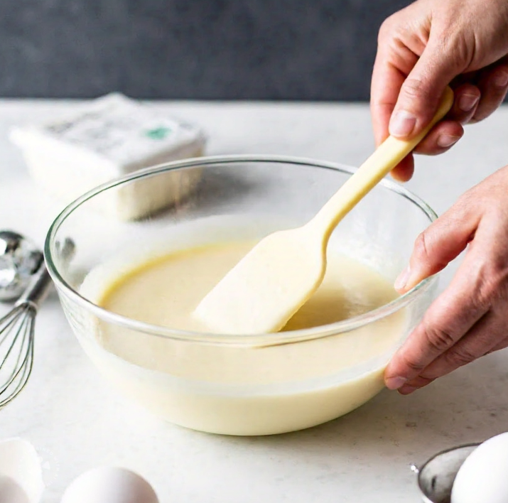 A baker mixing cheesecake batter in a glass bowl with no flour.