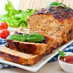 Perfectly smoked meatloaf on a wooden cutting board with a caramelized glaze, surrounded by fresh herbs and a smoking grill in the background.