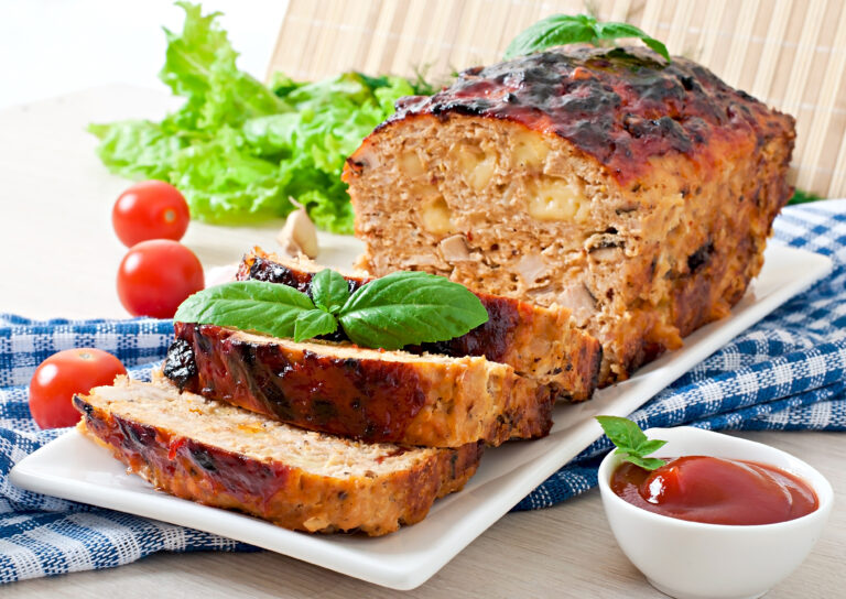 Perfectly smoked meatloaf on a wooden cutting board with a caramelized glaze, surrounded by fresh herbs and a smoking grill in the background.