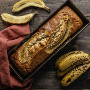 A freshly baked loaf of 4 ingredient banana bread on a wooden cutting board, surrounded by ripe bananas and a knife for slicing.