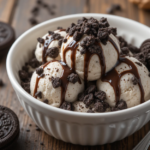 A close-up of a bowl of Oreo ice cream topped with crushed Oreos and a drizzle of chocolate syrup, served with a spoon.