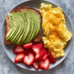 Scrambled eggs served with strawberries and avocado toast on a plate
